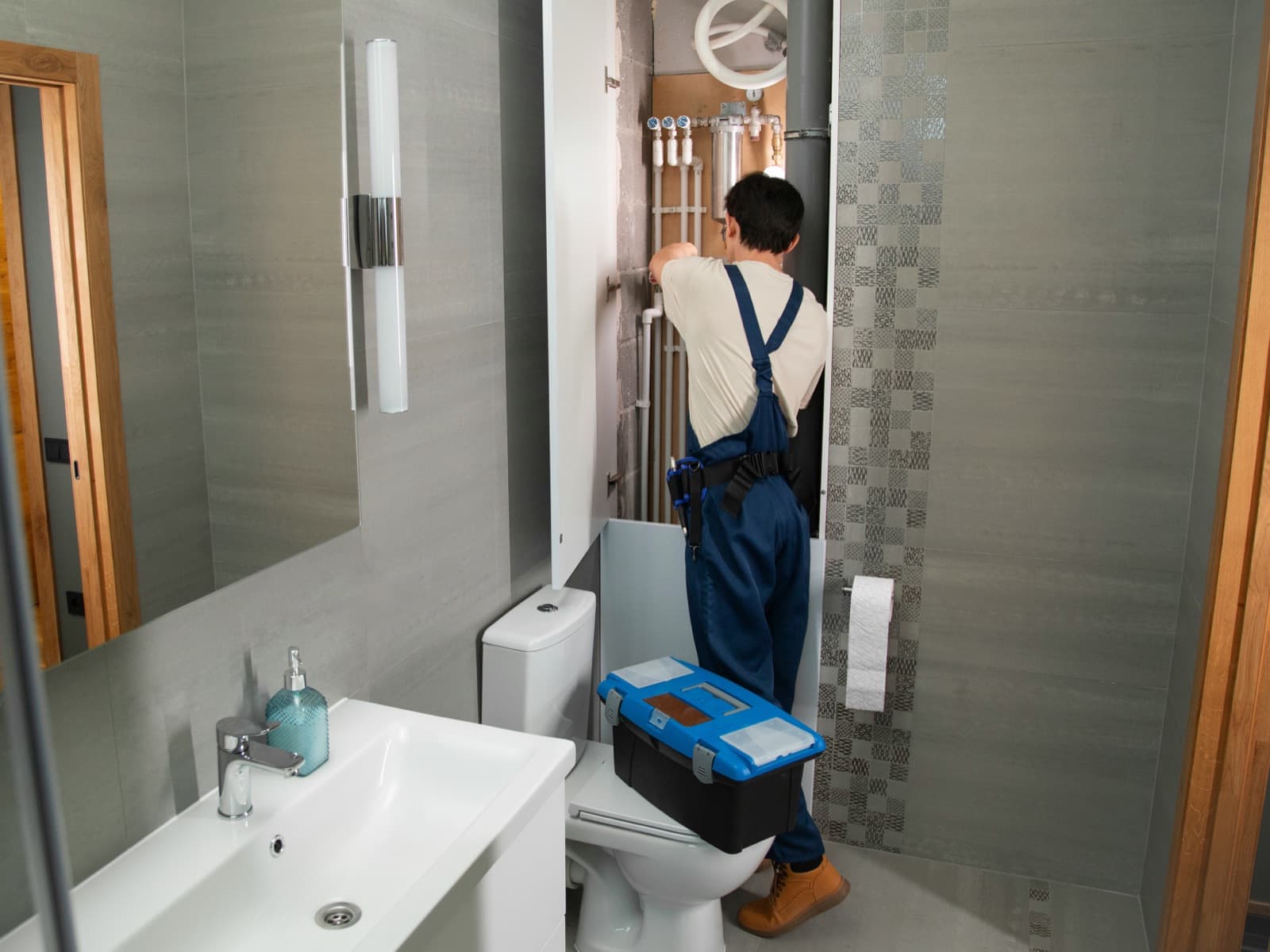 Plumber in blue overalls working on wall-mounted water lines in a tiled bathroom